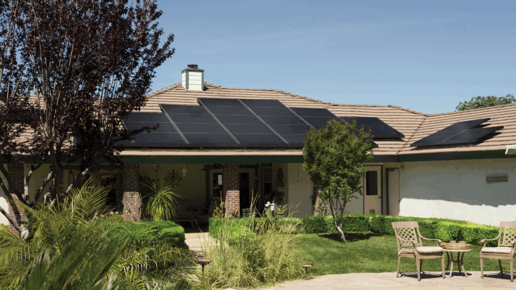 The picture shows a suburban home with a pale colour roof that has dark navy solar panels on top. The house has a small grass lawn at the front with some trees and greenery. There are 2 outdoor chaires and a table on the bottom right corner of the picture.